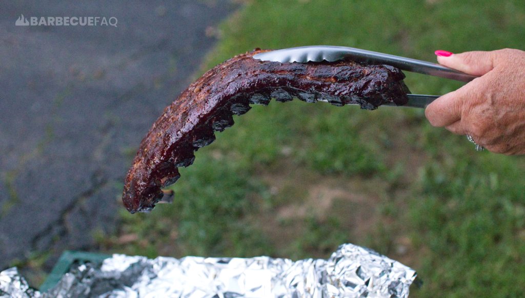 A person holds a rack of smoked ribs with tongs, performing a bend test to check tenderness. The ribs have a dark, caramelized crust and are curved, indicating they are cooked to a tender, juicy consistency