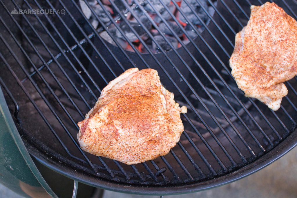 Two seasoned chicken thighs placed on a grill over indirect heat, with coals glowing beneath the grate. The meat is coated in a dry rub, ready to be smoked or grilled to develop a flavorful crust