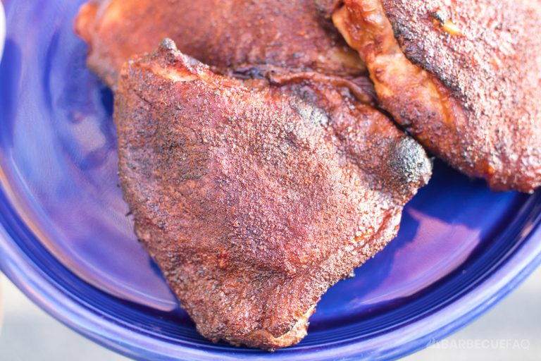 Close-up of smoked chicken thighs with a rich, dark crust and a seasoned, crispy skin, served on a vibrant blue plate. The smoky, charred surface suggests a flavorful, tender interior