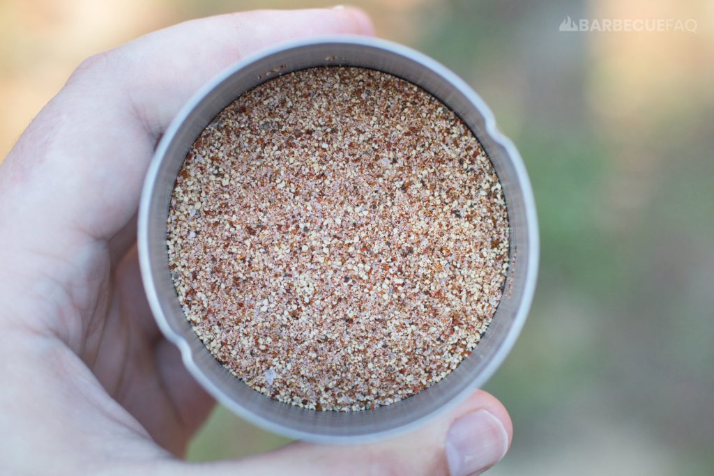 Close-up of a hand holding a metal container filled with a sweet and smoky BBQ rub, featuring a coarse blend of spices in shades of red, brown, and off-white, suggesting a balanced mix of salt, pepper, and other seasonings