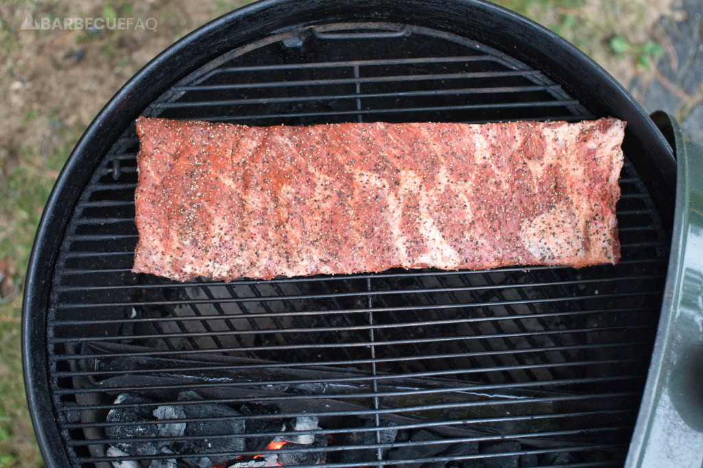 Rack of seasoned Texas-style spare ribs placed on a smoker grill over indirect heat, with charcoal and wood below to infuse a smoky flavor. The ribs have a dry rub coating, ready for slow smoking