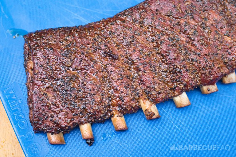 Rack of Texas-style smoked spare ribs with a dark, peppery crust and a glossy, caramelized surface, resting on a blue cutting board. The ribs have a rich, smoky appearance with visible seasoning, highlighting their savory flavor