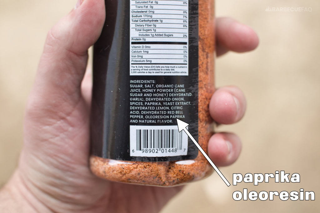 person holding a dry rub shaker bottle showing paprika oleoresin in ingredients list